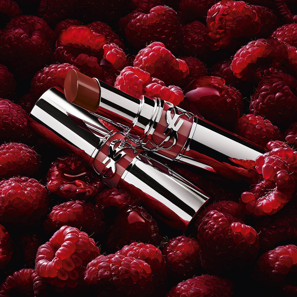 Two YSL Loveshine lipsticks, one open and one closed, in Berry Crush shade, nestled among fresh raspberries.