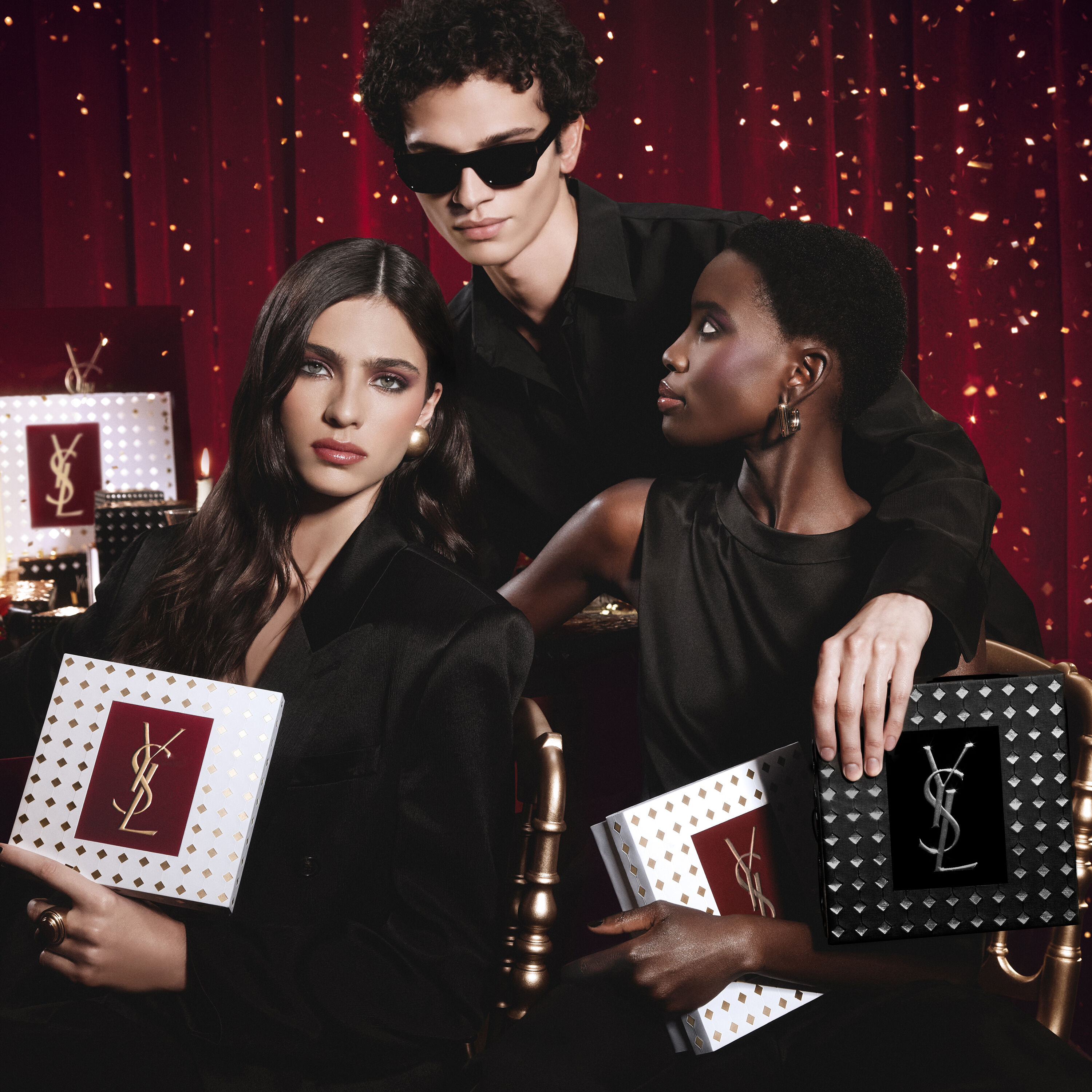 Model wearing a black suit and glam look, seated in a festive candlelit setting with YSL gift boxes.