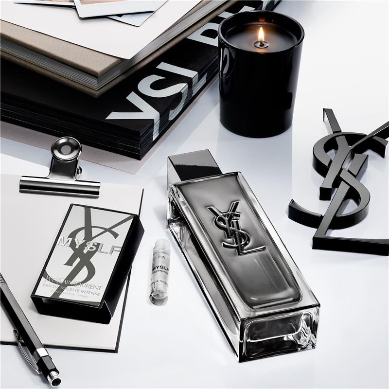 MYSLF Eau de Toilette Intense sample vial placed next to the full bottle in a sleek workspace with YSL notebooks and candle.